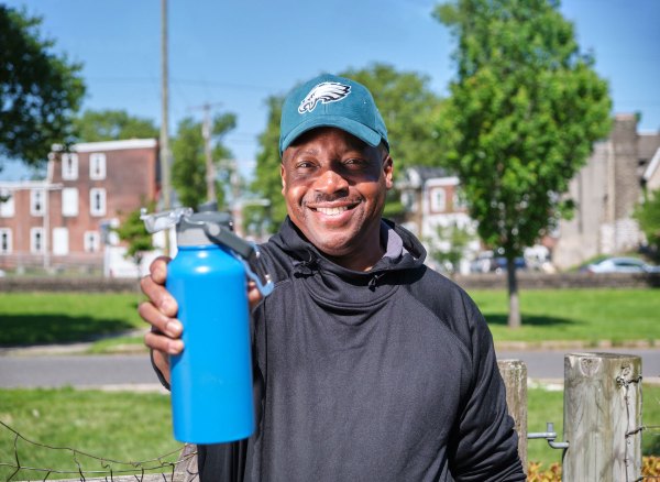 photo of Michael wearing an Eagles cap, standing in his neighborhood's community garden, holding out his reusable bottle of Philly tap water, with Philly rowhomes in the background