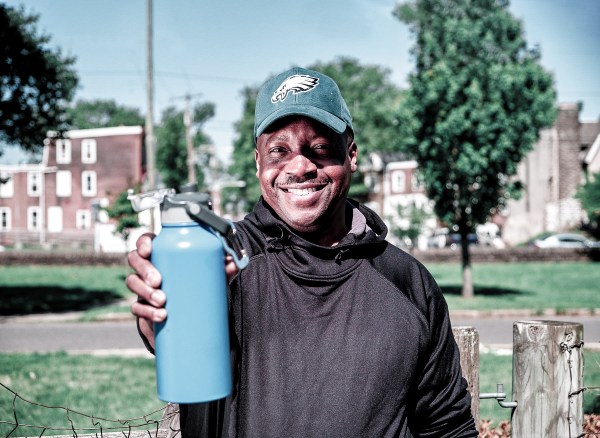 photo of Michael wearing an Eagles cap, standing in his neighborhood's community garden, holding out his reusable bottle of Philly tap water, with Philly rowhomes in the background
